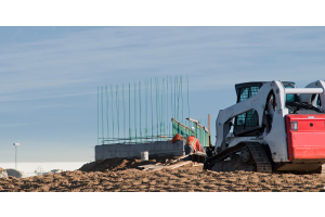 Bobcat compact loader working on a jobsite with workers and cement foundations in background