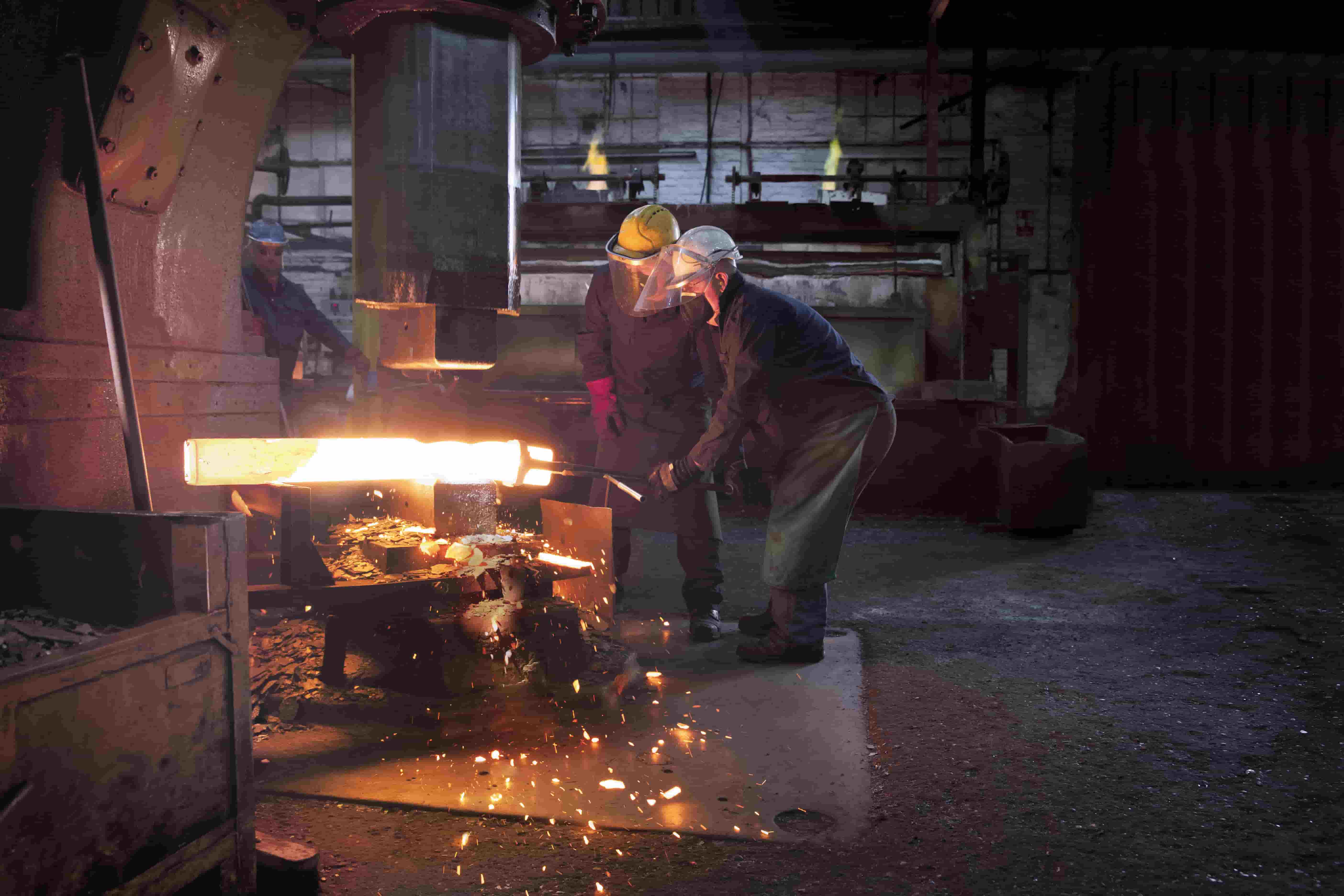 Two workers working a molten billet of metal with a power hammer